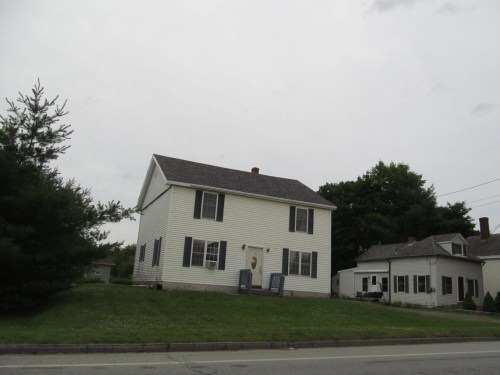 A two-story white house with black shutters, set on a grassy lot, surrounded by trees and other buildings under a cloudy sky.