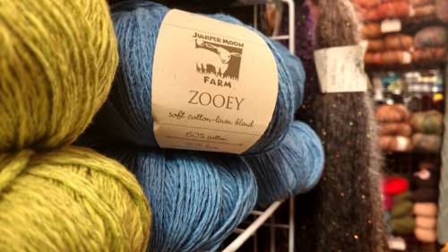 Skeins of colorful yarn in a store, featuring a label for "Zooey" by Juniper Moon Farm, a cotton-linen blend.