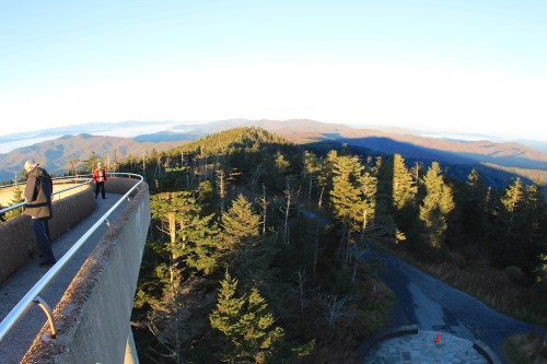 A scenic view from a mountaintop overlook, featuring trees and distant mountains under a clear sky. Two people stand nearby.