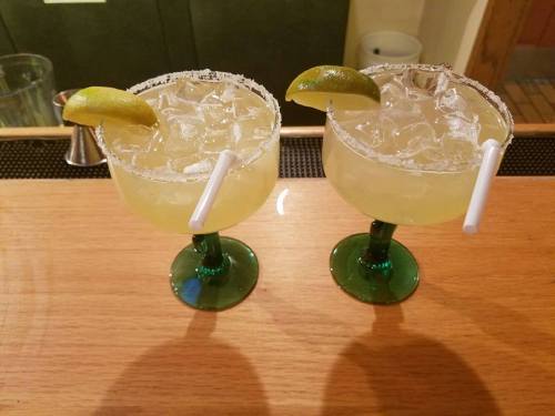 Two margarita glasses with lime wedges, ice, and salt rims, placed on a wooden bar counter.