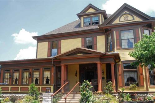 A large, colorful Victorian-style house with a porch, surrounded by greenery and blue skies.