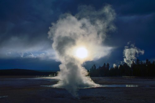 A dramatic scene of steam rising from geothermal features under a moody sky, with the sun partially obscured by clouds.