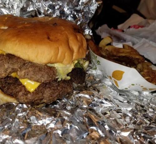 A close-up of a cheeseburger with two patties, lettuce, and cheese, wrapped in foil, alongside a portion of fries.