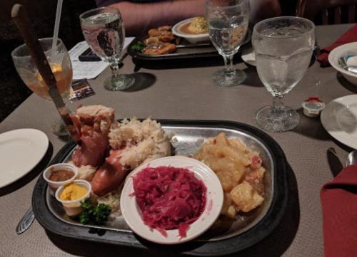 A metal platter with a large piece of meat, sauerkraut, red cabbage, and potatoes, accompanied by sauces and drinks.