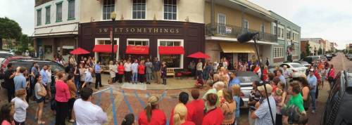 A large crowd gathers outside a bakery called "Sweet Somethings," with red umbrellas and a festive atmosphere.