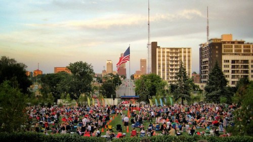A large crowd gathers on a grassy area for an outdoor event, with a U.S. flag and city skyline in the background.