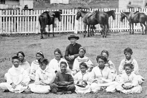 A historical black-and-white photo of a group of women and children seated outdoors, with horses in the background.