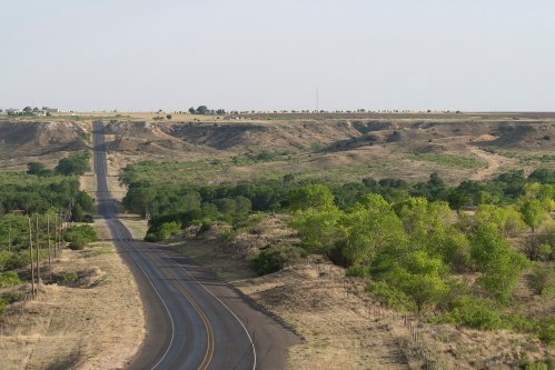 A winding road stretches through a hilly landscape with sparse trees and open fields under a clear sky.