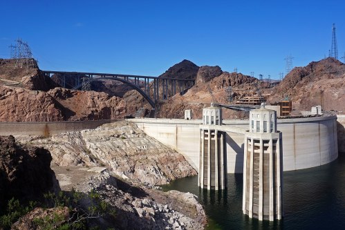 A large dam with two concrete towers, surrounded by rocky terrain and a clear blue sky. Power lines stretch in the background.