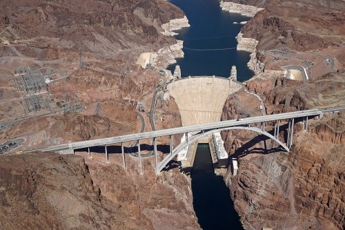 Aerial view of a large dam surrounded by rocky terrain and a reservoir, with a bridge spanning across the canyon.