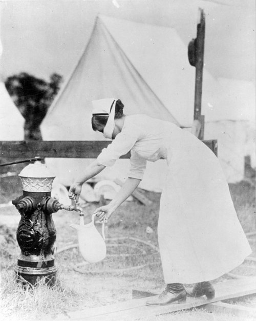 A nurse in historical attire fills a pitcher from a fire hydrant near tents in a camp setting.