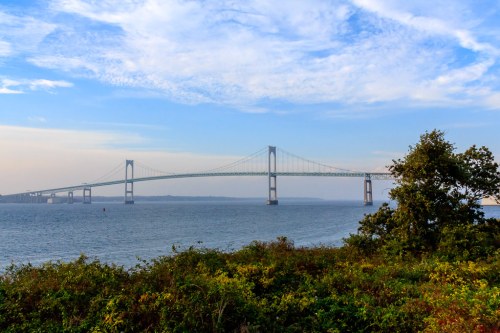 A suspension bridge spans a calm body of water, framed by greenery and a blue sky with scattered clouds.