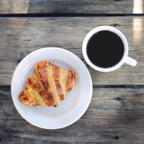 A golden croissant on a white plate next to a cup of black coffee on a wooden table.