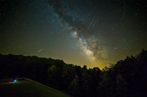 A stunning night sky filled with stars and the Milky Way, with shooting stars and a faint glow from the horizon.