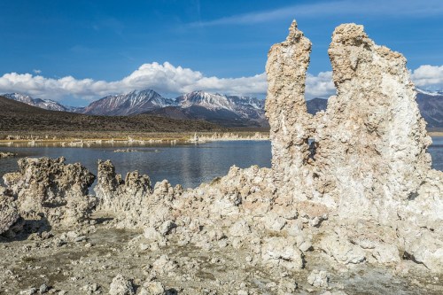 Rock formations rise from the shore of a lake, with mountains and blue sky in the background.