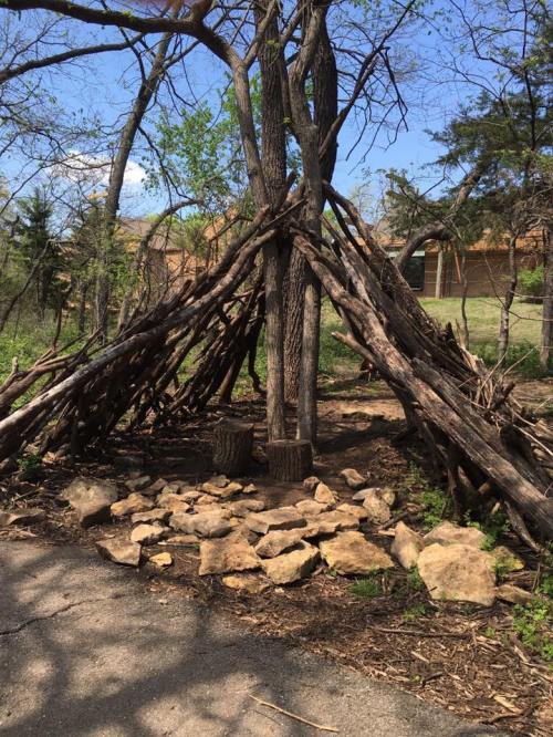 A rustic structure made of branches and stones, surrounded by trees and greenery, in a natural outdoor setting.