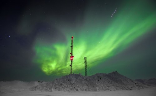 A vibrant green aurora borealis illuminates the night sky above communication towers and snow-covered terrain.