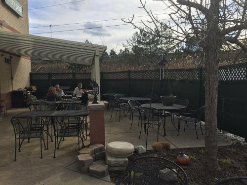 Outdoor dining area with empty tables, a few patrons, and a tree, set against a backdrop of greenery and a cloudy sky.