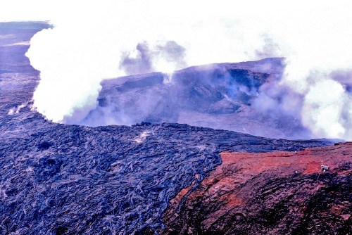 Aerial view of a volcanic landscape with smoke rising from fissures and dark lava flows contrasting with lighter terrain.