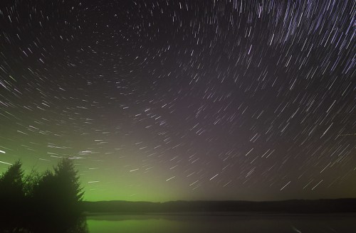 Star trails swirl over a calm lake, with a greenish glow on the horizon and silhouettes of trees in the foreground.