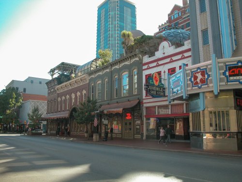 Historic buildings line a city street, featuring shops and a movie theater, with modern skyscrapers in the background.