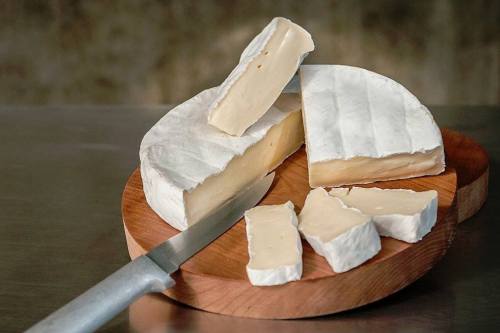 A round wheel of soft cheese with a white rind, partially sliced, on a wooden cutting board with a knife.