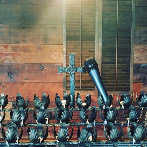 A microphone stands next to a decorative cross in front of a wooden backdrop with shutters.