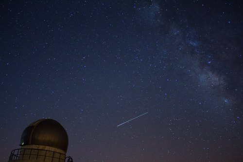 A starry night sky with the Milky Way and a bright meteor streaking across, above a dome-shaped observatory.