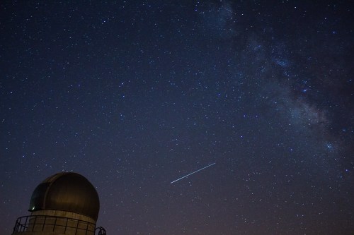 A starry night sky with the Milky Way and a bright meteor streaking across, above a dome-shaped observatory.