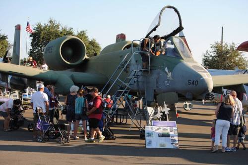 A crowd gathers around a military aircraft at an outdoor event, with people exploring and taking photos.