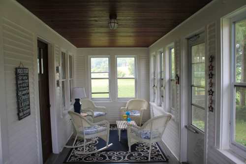 A cozy sunroom with white wicker furniture, a patterned rug, and large windows overlooking a green landscape.