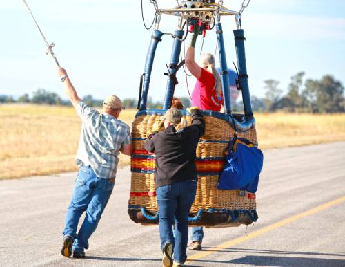 Three people assist in lifting a colorful hot air balloon basket onto a runway under a clear blue sky.