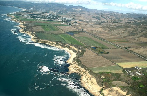 Aerial view of a coastal landscape featuring cliffs, farmland, and the ocean with waves crashing against the shore.