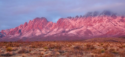 Vibrant pink-hued mountains under a cloudy sky, surrounded by a desert landscape with sparse vegetation.
