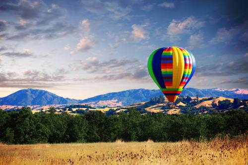 A colorful hot air balloon floats over a scenic landscape of rolling hills and trees under a blue sky.