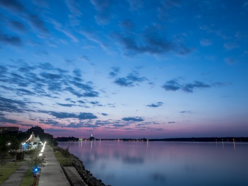 A serene riverside view at dusk, with a colorful sky reflecting on calm waters and a pathway lined with lights.