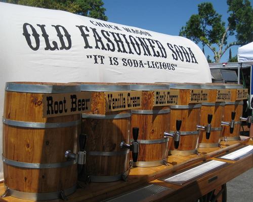 A wooden soda fountain with various flavors like root beer and vanilla cream, under a sign that reads "Old Fashioned Soda."