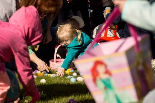 A young child in a teal jacket reaches for colorful Easter eggs on the grass, with adults and baskets nearby.