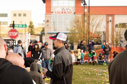 A crowd gathers at an outdoor event, with children and adults participating in an egg hunt near a building.