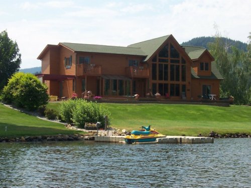 A large wooden house by a lake, featuring a green lawn, colorful flowers, and a small dock with a yellow watercraft.