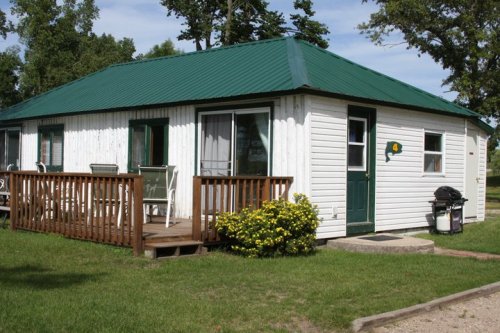 A small white cottage with a green metal roof, wooden deck, and yellow flowers in front, set in a grassy area.