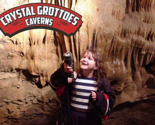 A smiling child holds a flashlight in a cave, with rock formations in the background and a "Crystal Grottoes Caverns" sign.