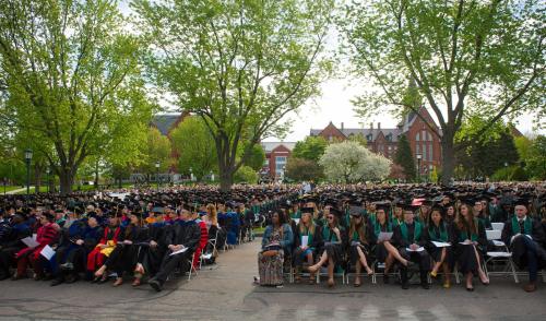 A large crowd of graduates in caps and gowns seated outdoors, surrounded by trees and historic buildings during a ceremony.