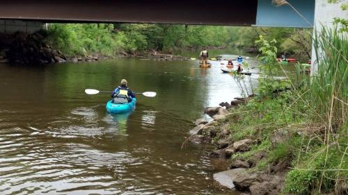 A group of kayakers paddles down a calm river under a bridge, surrounded by lush greenery.