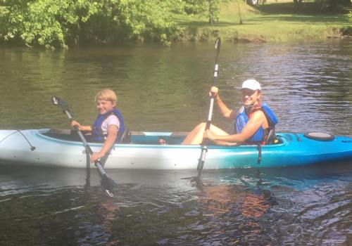 A woman and a child paddle together in a kayak on a calm river surrounded by greenery.