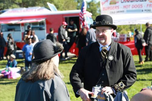 A man in a top hat and suit talks to a woman at a food truck event, with people and food trucks in the background.