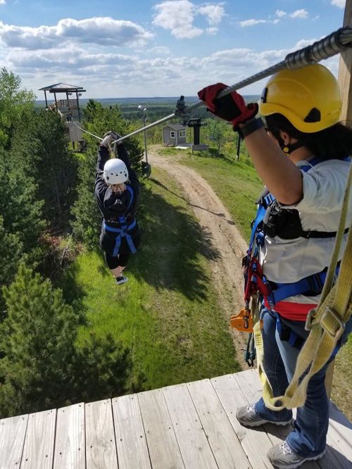 A person zip-lining from a platform while another person assists from the side, surrounded by trees and a blue sky.
