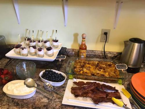 A breakfast spread featuring yogurt parfaits, a casserole, bacon, fresh strawberries, blueberries, and hot sauce.