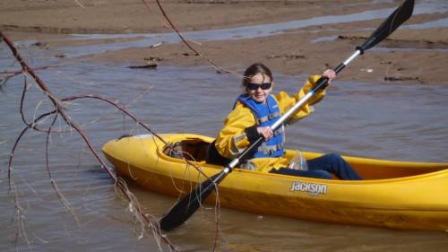A person in a yellow kayak paddles through muddy water, wearing a blue life jacket and sunglasses.