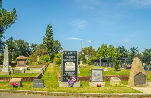 A peaceful cemetery scene with gravestones, greenery, and a small American flag at a grave. Clear blue sky above.